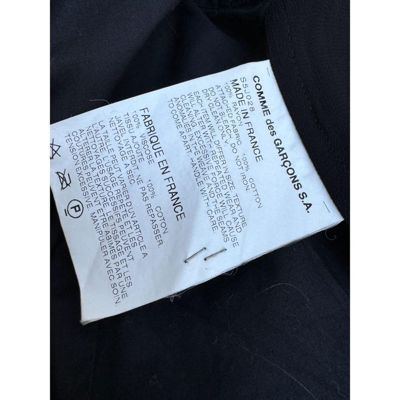 Pode incluir: Uma etiqueta branca com texto preto da Comme des Gar&ccedil;ons S.A. est&aacute; presa a um tecido preto. A etiqueta diz "Made in France" e inclui a composi&ccedil;&atilde;o do tecido e as instru&ccedil;&otilde;es de cuidado. A etiqueta &eacute; fixada com dois grampos.