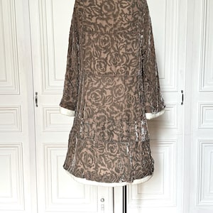 Puede incluir: Un abrigo de terciopelo marr&oacute;n con un estampado de rosas, exhibido en un maniqu&iacute;. El abrigo tiene un cuello ancho, mangas largas y una falda escalonada. Ribetes blancos acent&uacute;an los pu&ntilde;os y el dobladillo. El abrigo es de estilo vintage.