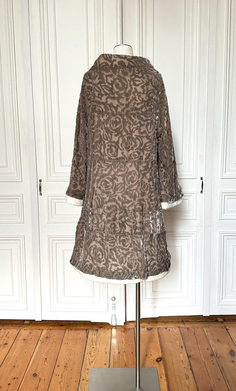 Puede incluir: Un abrigo de terciopelo marr&oacute;n con un estampado de rosas, exhibido en un maniqu&iacute;. El abrigo tiene un cuello ancho, mangas largas y una falda escalonada. Ribetes blancos acent&uacute;an los pu&ntilde;os y el dobladillo. El abrigo es de estilo vintage.