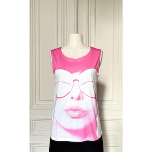 May include: A sleeveless white and pink top featuring a graphic of a face with sunglasses and a kiss. The top has a pink neckline and shoulder straps. The graphic is in shades of pink and white.