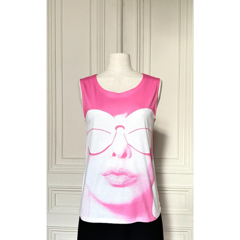 May include: A sleeveless white and pink top featuring a graphic of a face with sunglasses and a kiss. The top has a pink neckline and shoulder straps. The graphic is in shades of pink and white.