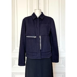 May include: A dark navy blue jacket featuring a classic collar, long sleeves, and a concealed front button placket. The garment includes a horizontal zippered pocket on the right chest and a vertical zippered pocket on the left side of the torso.