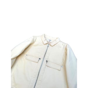 May include: A cream-colored zip-up jacket with a collar and two large pockets on the front. The jacket has a zipper closure and a small label on the collar that says "Collaborations" in black text.