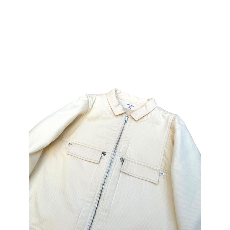 May include: A cream-colored zip-up jacket with a collar and two large pockets on the front. The jacket has a zipper closure and a small label on the collar that says "Collaborations" in black text.