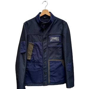 Peut inclure: Une veste utilitaire bleu marine dotée d'un col montant, d'une fermeture à boutons sur le devant et de plusieurs empiècements contrastants bleu foncé et vert olive. La veste comprend une poche poitrine avec fenêtre transparente.