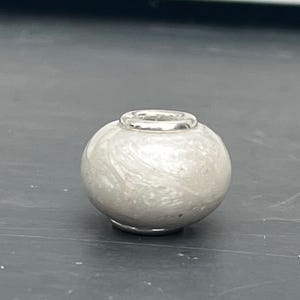 Op de afbeelding: Een witte glazen kraal met een zilveren metalen band om het midden. De kraal heeft een marmereffect.
