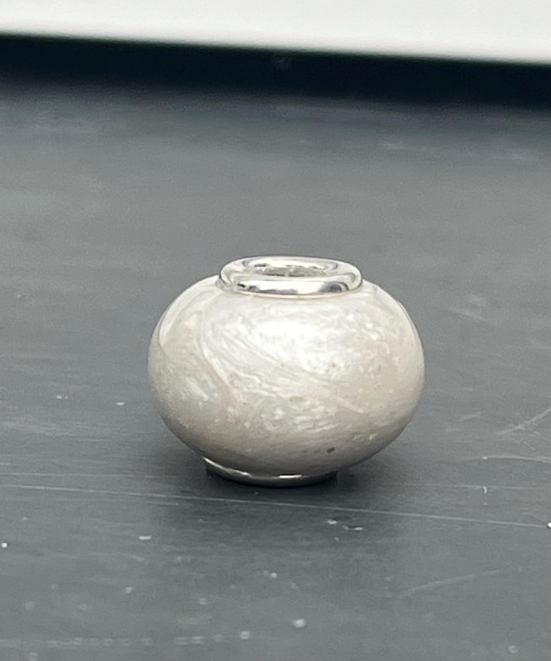Op de afbeelding: Een witte glazen kraal met een zilveren metalen band om het midden. De kraal heeft een marmereffect.
