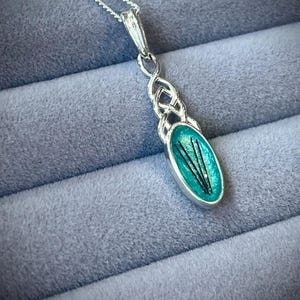 Op de afbeelding: Zilveren hangerketting met een ovaal turquoise geëmailleerd middenstuk en een zwart lineair ontwerp. De hanger heeft een Keltisch knoopontwerp en hangt aan een fijne zilveren ketting. De ketting wordt gepresenteerd op een grijze fluwelen ondergrond.