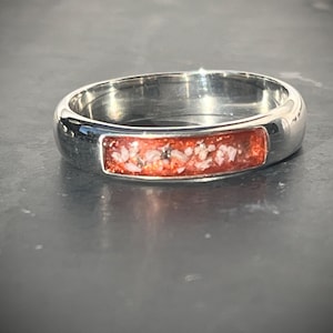 Op de afbeelding: Een ring van gepolijst zilverkleurig metaal met een rechthoekige inleg. De inleg bevat een feloranje harsbasis met kleine, onregelmatige witte en donkergrijze spikkels, wat zorgt voor een textuur in de gladde metalen band.
