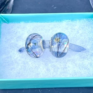 Op de afbeelding: Twee heldere glazen kralen met zwarte en zilveren accenten. De kralen bevinden zich in een lichtblauwe doos met witte voering.