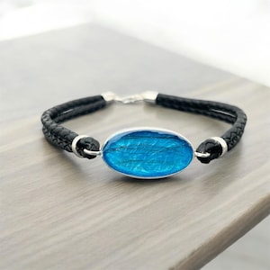 Op de afbeelding: Een zwart leren armband met een zilveren ovale bedel. De bedel is gevuld met blauwe hars en bevat ingebed haar.