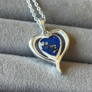 Op de afbeelding: Een zilveren ketting met een hartvormige hanger. De hanger heeft een dubbel hart, met een blauw centrum met witte en zwarte vlekken. De ketting is ook zilver.