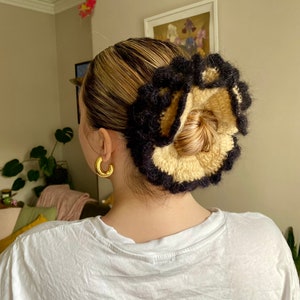 May include: A black and beige crocheted hair scrunchie with a ruffled design. The scrunchie is worn in a woman's hair.