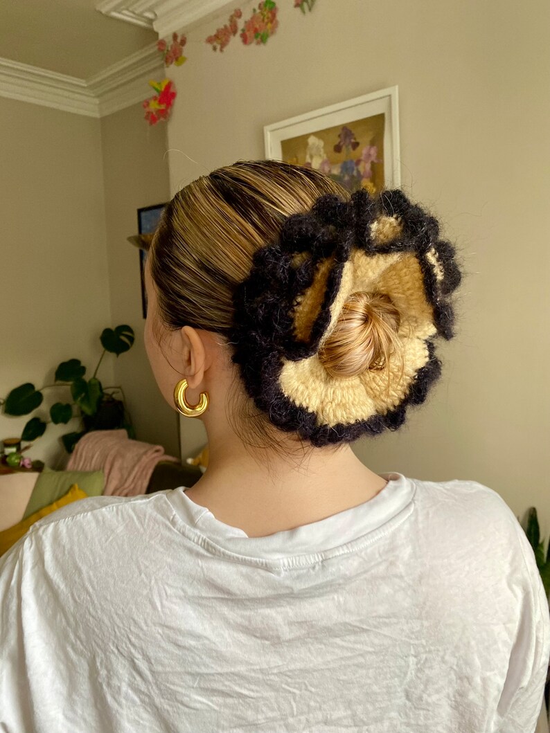 May include: A black and beige crocheted hair scrunchie with a ruffled design. The scrunchie is worn in a woman's hair.