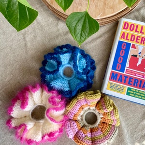 May include: Three crocheted hair scrunchies in different colours and patterns. One is blue, one is white with pink accents, and one is multi-coloured with pink, orange, and green.