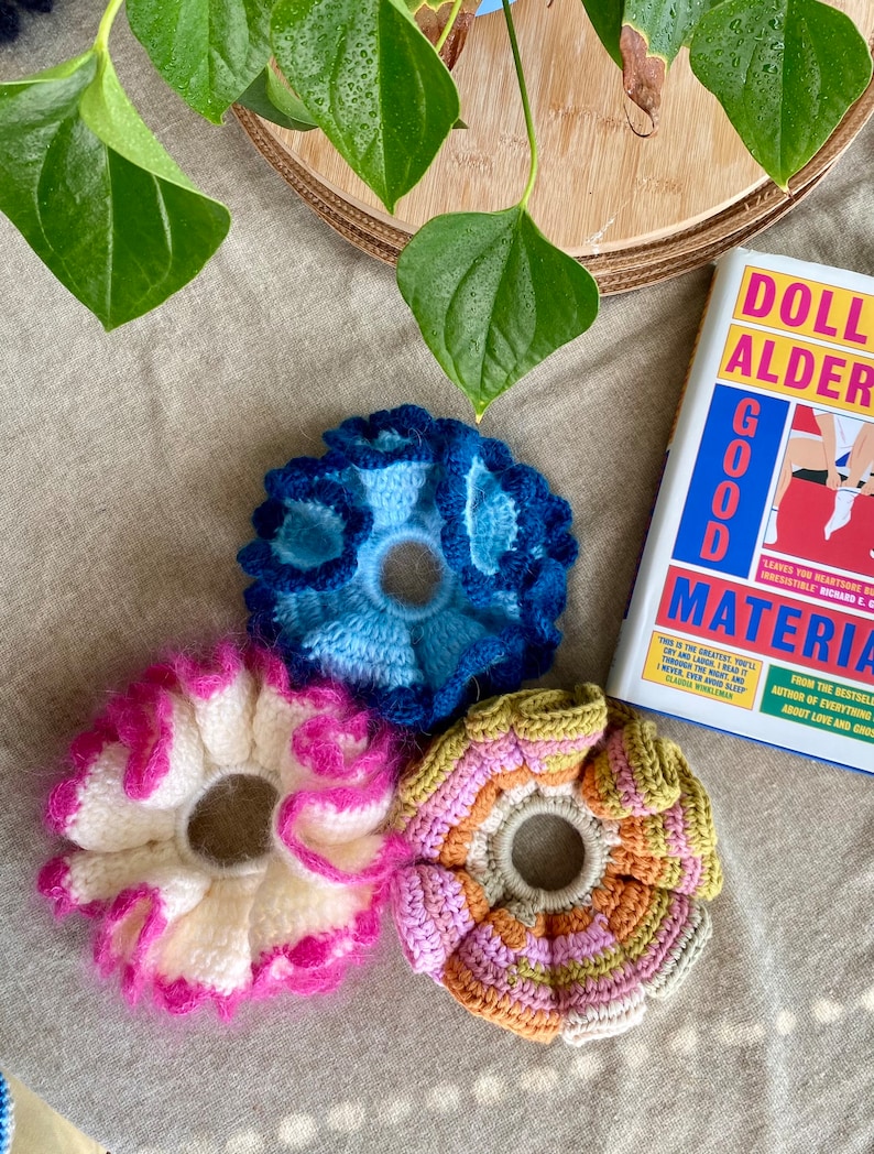 May include: Three crocheted hair scrunchies in different colours and patterns. One is blue, one is white with pink accents, and one is multi-coloured with pink, orange, and green.