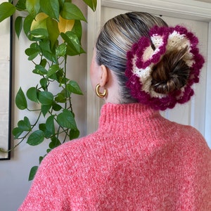 May include: A pink and white crocheted hair scrunchie with a ruffled design. The scrunchie is worn in a woman's hair, pulled back into a bun.