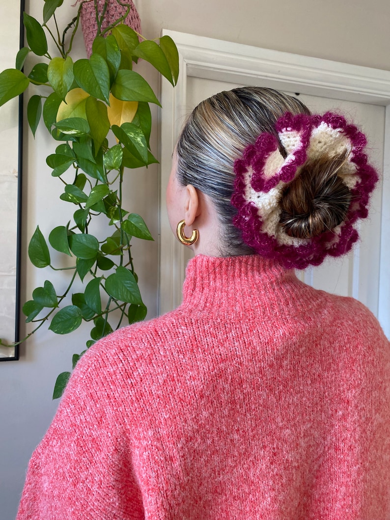 May include: A pink and white crocheted hair scrunchie with a ruffled design. The scrunchie is worn in a woman's hair, pulled back into a bun.