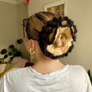 May include: A close-up of a woman's hair styled in a bun with a black and beige crocheted hair scrunchie. The scrunchie is in the shape of a flower with a ruffled edge.