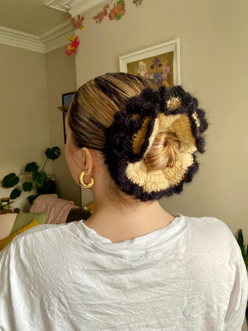 May include: A close-up of a woman's hair styled in a bun with a black and beige crocheted hair scrunchie. The scrunchie is in the shape of a flower with a ruffled edge.
