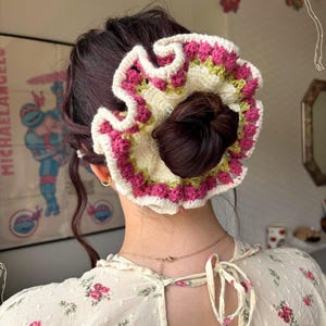 Può includere: Un elastico per capelli all'uncinetto fatto a mano in crema, rosa e verde. L'elastico è circolare con un bordo a balze e viene indossato in uno chignon. Questo accessorio per capelli è un'aggiunta elegante a qualsiasi acconciatura.
