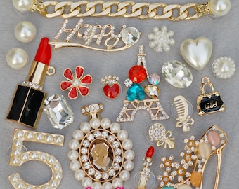 May include: Assortment of charms for shoes or bags, featuring a variety of designs including a lipstick, a perfume bottle, a Eiffel Tower, a shoe, a number 5, a flower, a heart, a purse, a comb, a snowflake, and a chain link. The charms are made of metal and decorated with rhinestones and pearls.