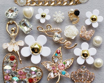 May include: A collection of decorative charms in various shapes and designs. Includes a gold chain, flowers, hearts, a ballerina, and a crown, all adorned with rhinestones, pearls, and crystals. The charms are in gold, white, and clear colors.