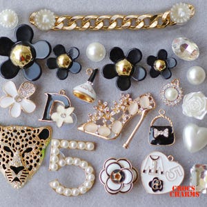May include: A collection of black and white charms for shoes, bags, or other accessories. The charms include a leopard, a shoe, a purse, a flower, a number 5, a letter D, and a Chanel-inspired logo. The charms are made of metal, plastic, and pearls.