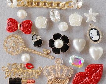 May include: A collection of decorative charms in various shapes and colors. Includes a red bow, a black flower with a pearl center, a gold key, a crown, and a pair of red lips. The charms are embellished with rhinestones and pearls.