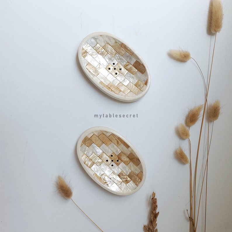 Puede incluir: Dos jaboneras ovaladas blancas con un dise&ntilde;o de mosaico dorado y blanco. Las jaboneras tienen agujeros de drenaje.