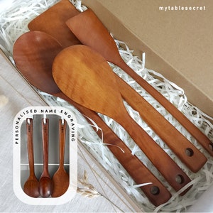 May include: Four wooden kitchen utensils in a box with white shredded paper. The utensils include a spatula, a large spoon, and two smaller spoons. The box has a label that says "Personalised Name Engraving".