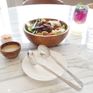 May include: A wooden bowl filled with a salad containing grilled meat, vegetables, and potatoes. A white plate with two silver serving tongs is on the table.