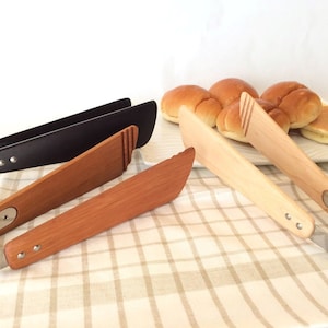 Puede incluir: Dos pares de pinzas de cocina, una negra y otra de madera, con mango de acero inoxidable. Las pinzas de madera están sobre un mantel a cuadros blanco y marrón. También hay algunos panecillos y una tortita en un plato blanco en el fondo.