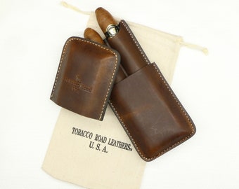 Puede incluir: Dos estuches de cuero marrón para cigarros con una bolsa de algodón blanco con cordón. La bolsa tiene el texto "Tobacco Road Leathers, U.S.A." impreso en ella.