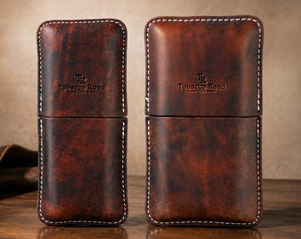 May include: Two rectangular brown leather cigar cases with visible white stitching along the edges. Each case features a debossed logo with the text Tobacco Road USA. The leather has a dark, mottled, and burnished finish with a smooth texture.