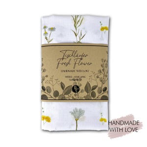 Puede incluir: Un camino de mesa blanco plegado con diseños florales y el texto "Tischläufer Fresh Flower". El camino de mesa está envuelto con una banda de papel marrón que también tiene texto e ilustraciones florales. El camino de mesa tiene flores amarillas y blancas.