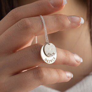 Puede incluir: Collar de plata con dos colgantes redondos, uno con el nombre "Olivia" y el otro con el nombre "Jack".