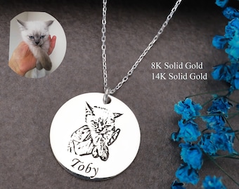 May include: A silver necklace with a round pendant featuring a black and white engraved image of a cat with the name "Toby" underneath. The necklace is on a white background with blue flowers.
