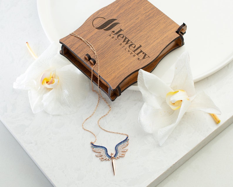 May include: A rose gold necklace with a pendant shaped like angel wings, featuring blue and white gemstones. The necklace is displayed with a wooden jewelry box and white orchids. The box has the text "Jewelry Art Silver".