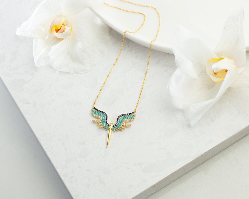May include: A gold-colored necklace with a pendant shaped like angel wings. The wings are adorned with blue, turquoise, and clear gemstones. The necklace is displayed on a white surface, with white flowers in the background.