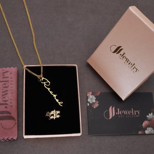 May include: A gold-colored necklace with the name "Rachel" in a cursive font, displayed in a black box. The box is inside a pink jewelry box. A pink cloth and a black business card with the brand logo are also visible.