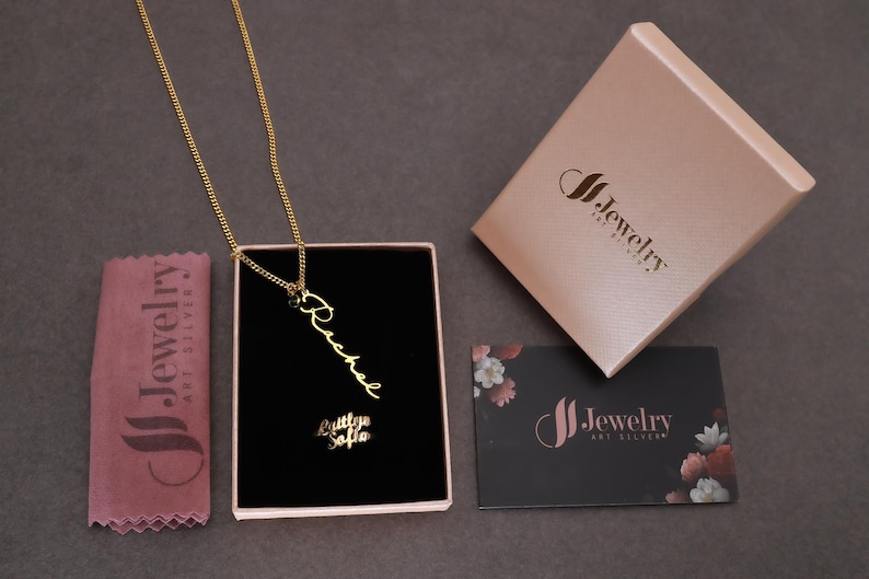 May include: A gold-colored necklace with the name "Rachel" in a cursive font, displayed in a black box. The box is inside a pink jewelry box. A pink cloth and a black business card with the brand logo are also visible.