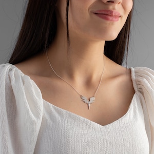 May include: A silver necklace featuring an angel wing pendant with a central, pointed design. The pendant is adorned with small, sparkling embellishments. The necklace is displayed on a person wearing a white, textured top.