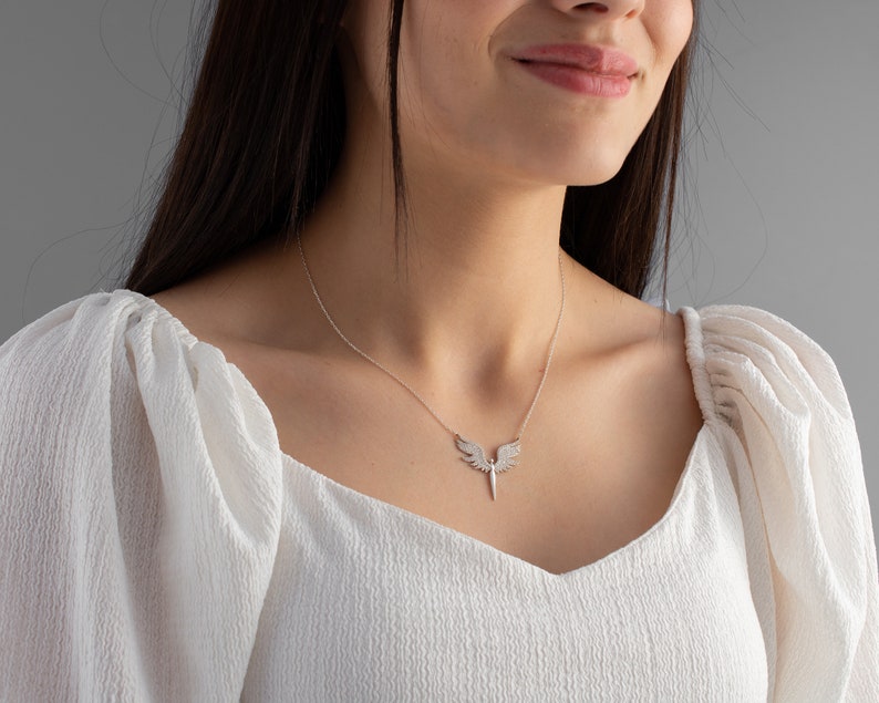 May include: A silver necklace featuring an angel wing pendant with a central, pointed design. The pendant is adorned with small, sparkling embellishments. The necklace is displayed on a person wearing a white, textured top.