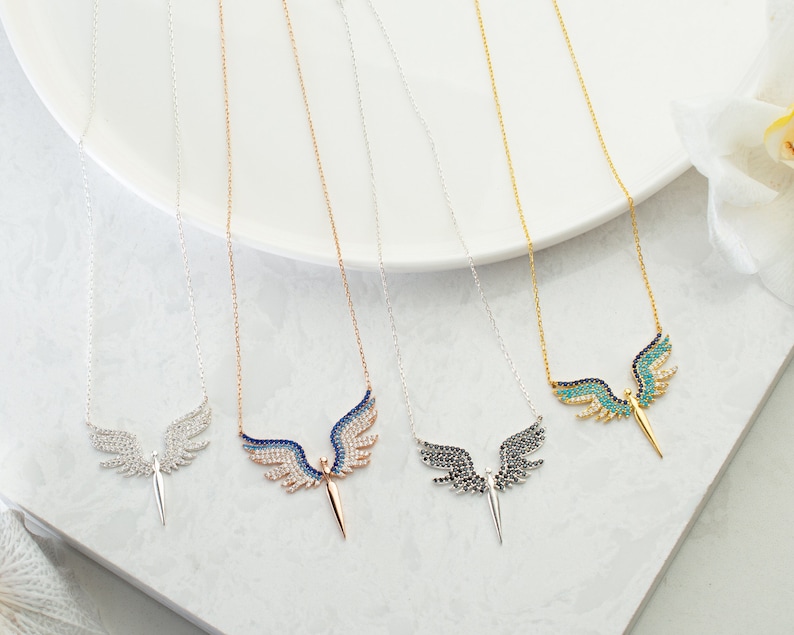 May include: A collection of angel wing necklaces in silver, rose gold, and gold tones. Each necklace features a pendant with detailed wings and a central, pointed element. The wings are adorned with various colored gemstones.