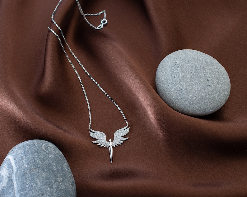 May include: A silver necklace with a pendant shaped like angel wings and a sword. The wings are encrusted with small, sparkling stones. The necklace rests on a brown satin fabric, with two gray stones in the background.