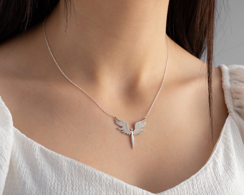 May include: A silver necklace featuring a pendant shaped like angel wings with a central, pointed element. The wings are encrusted with small, sparkling stones. The necklace is displayed on a person wearing a white top.