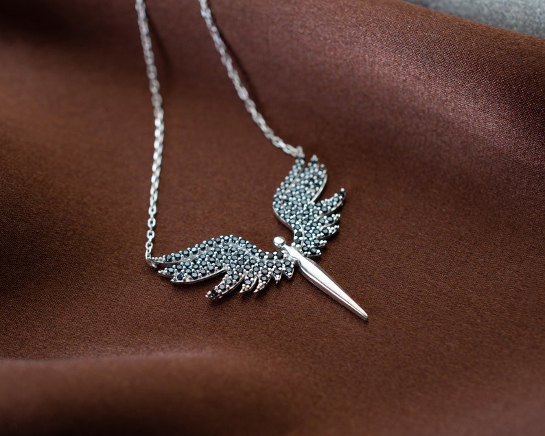 May include: A silver-toned necklace featuring an angel wing design. The wings are adorned with black gemstones, and the pendant has a pointed center. The necklace is displayed on a brown fabric background.