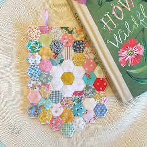 Może przedstawiać: Ręcznie robiony podkładka pod kubek z kwiatowym wzorem w kształcie plastra miodu. Podkładka ma centralny kształt kwiatu z żółtym środkiem, otoczony różnymi kolorowymi sześciokątnymi kawałkami materiału. Obok leży książka z zieloną okładką i kwiatową ilustracją.