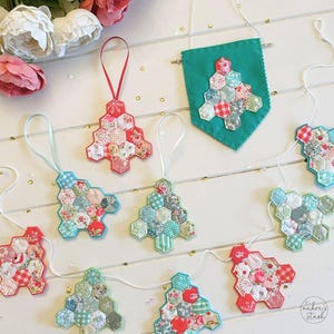 May include: A collection of handmade fabric Christmas tree ornaments and a banner. The ornaments are made from patchwork fabric in various floral and gingham patterns, with red, blue, and green accents. Each tree has a ribbon for hanging. The banner is teal felt with a patchwork tree.